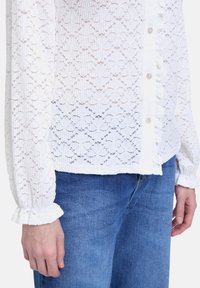 Camicia bianca in pizzo con bottoni, caratterizzata da polsini arricciati e un motivo floreale testurizzato, abbinata a jeans in denim blu.