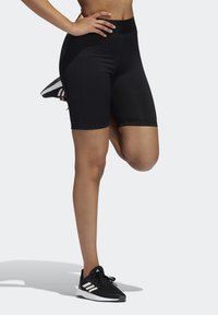 Shorts de sport noirs et extensibles avec une taille haute, présentant une texture lisse et une coupe ajustée, associés à des baskets noires et blanches.