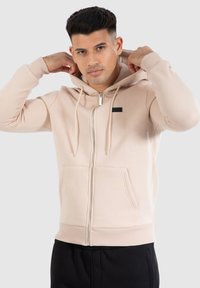 Haut de hoodie zippé beige clair avec une capuche, des poches avant, des poignets côtelés et un petit détail de label noir. Tissu doux avec une texture lisse.