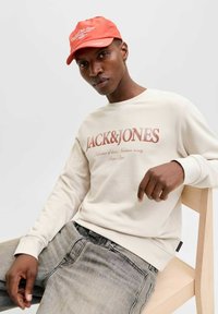 Sweat-shirt beige avec le texte imprimé "JACK&JONES" en rouge, associé à un jean gris clair. Le modèle porte une casquette rouge vif.