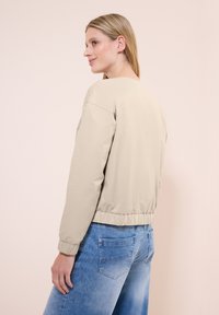 Beigefärgad bomberjacka med elastisk nederkant och manschetter, med en enkel design. Bärs med ljusblå jeans, sedd från sidan.