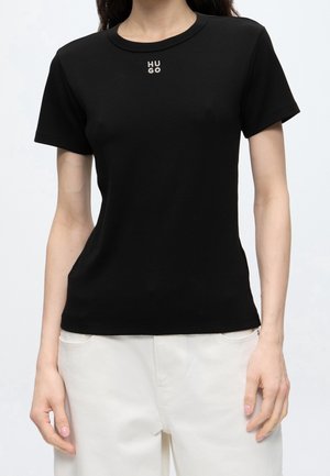 Femme portant un t-shirt noir cintré à manches courtes avec un petit texte blanc « "HUGO" » sur la poitrine, associé à un pantalon blanc taille haute.