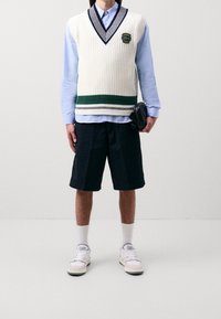 Biały dziergany kamizelka z zielonymi i granatowymi paskami, nałożona na jasnoniebieską koszulę. Czarna szorty i białe trampki dopełniają strój.