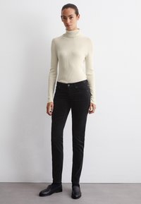 Un pull col roulé en crème à côtes, associé à un pantalon slim en velours côtelé noir, avec une fermeture à bouton et des poches latérales, porté avec des mocassins noirs.