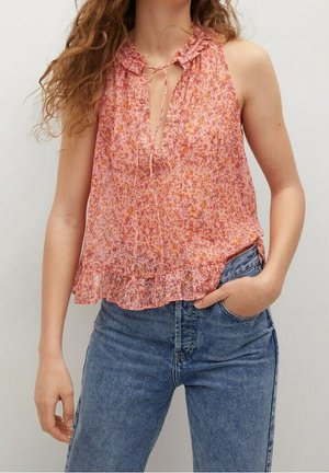 Mouwloze roze gebloemde blouse met gerimpelde kraag en zoom, strikdetail bij de halslijn, gedragen met een high-waisted blauwe jeans, hand in de zak.
