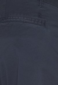 Tissu bleu foncé avec une poche passepoilée horizontale et un petit logo cousu au-dessus de l'ouverture de la poche.