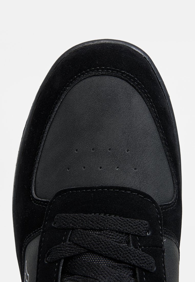 Zapatilla negra con parte superior de ante texturizado y cuero liso, puntera redondeada, detalles perforados y cordones planos.