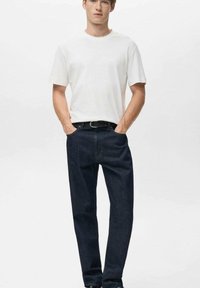 Mlad moški stoji z rokami v žepih, oblečen v enostavno belo majico s kratkimi rokavi in temno modre kavbojke z črnim pasom.