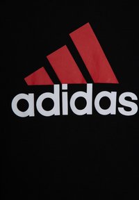 Camiseta negra con un gran logo de Adidas en rojo y blanco. El logo incluye tres franjas diagonales encima de la palabra "adidas".