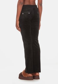 Pantalons évasés en velours noir avec une taille haute, deux poches arrières avec fermetures à pression, et une texture lisse. Portés avec des chaussons en fourrure marron.