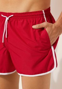 Rote Badeshorts aus leichtem Material, mit einem elastischen Bund und weißen Kordeln sowie kontrastierendem weißen Saum an den Kanten.