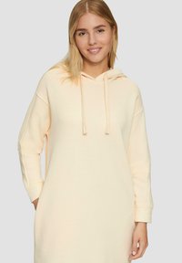 Helles Pfirsich-Hoodie-Kleid mit langen Ärmeln, Vordertaschen und Kordelzugkapuze. Weicher Stoff mit lockerer Passform und subtilen seitlichen Akzenten.