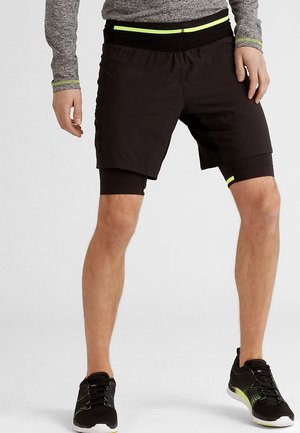 Person trägt schwarze, geschichtete Sportshorts mit neongelben Akzenten, schwarze Laufschuhe und ein graues Langarmoberteil, steht mit gespreizten Beinen.