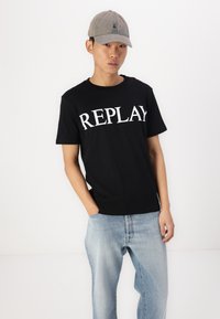 Camiseta negra con la palabra "REPLAY" en grandes letras blancas. Combinada con jeans de mezclilla azul claros. El modelo lleva una gorra gris con un logo.
