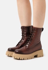 Botas de cuero marrón hasta el tobillo con suelas de goma beige gruesas, que cuentan con cordones en la parte frontal y una cremallera lateral para facilitar su uso. Parte superior texturizada y diseño resistente.
