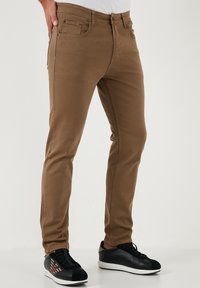 Smeđi hlače od pamuka s dodatkom, slim fit kroja, s pet džepova i suptilnim detaljima šivanja. Uparene s crnim tenisicama i bijelom košuljom.