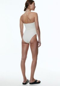 Maillot de bain une pièce blanc texturé avec un design à col halterneck. Jambes haute couture et finition lisse. Porté avec des sandales noires à enfiler.
