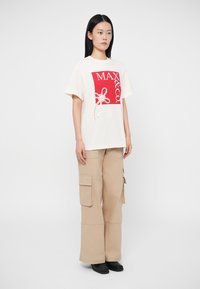 Νεαρή γυναίκα φοράει ένα λευκό υπερμεγέθες μπλουζάκι με κόκκινη εκτύπωση "MAX&CO." και μπεζ cargo παντελόνι, στέκεται μπροστά σε λευκό φόντο.