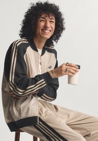 Beež ja must värvi sportlik dressipluus triibuliste aktsentidega, millel on lukuga jakk ja lõdvalt istuvad püksid; sile kangas ja võrgust detailid.