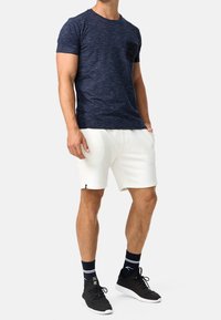 Man wearing navy blue short sleeve t-shirt, white shorts, black athletic shoes, and black crew socks against a plain white background.