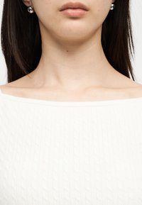 Κοντινό πλάνο του κάτω μέρους του προσώπου και του λαιμού μιας γυναίκας που φοράει ασημένια σκουλαρίκια σφαίρα και λευκό υφασμάτινο τοπ με τεχνουργήματα έξω από τους ώμους.