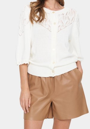 Kvinde iført hvid kortærmet strikket cardigan med knapper foran og beige højtaljede lædershorts, stående med den ene hånd i lommen.
