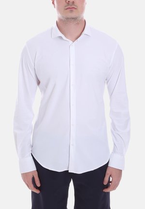 Uomo con indosso una camicia bianca a maniche lunghe con bottoni e pantaloni scuri, in piedi davanti a uno sfondo chiaro.