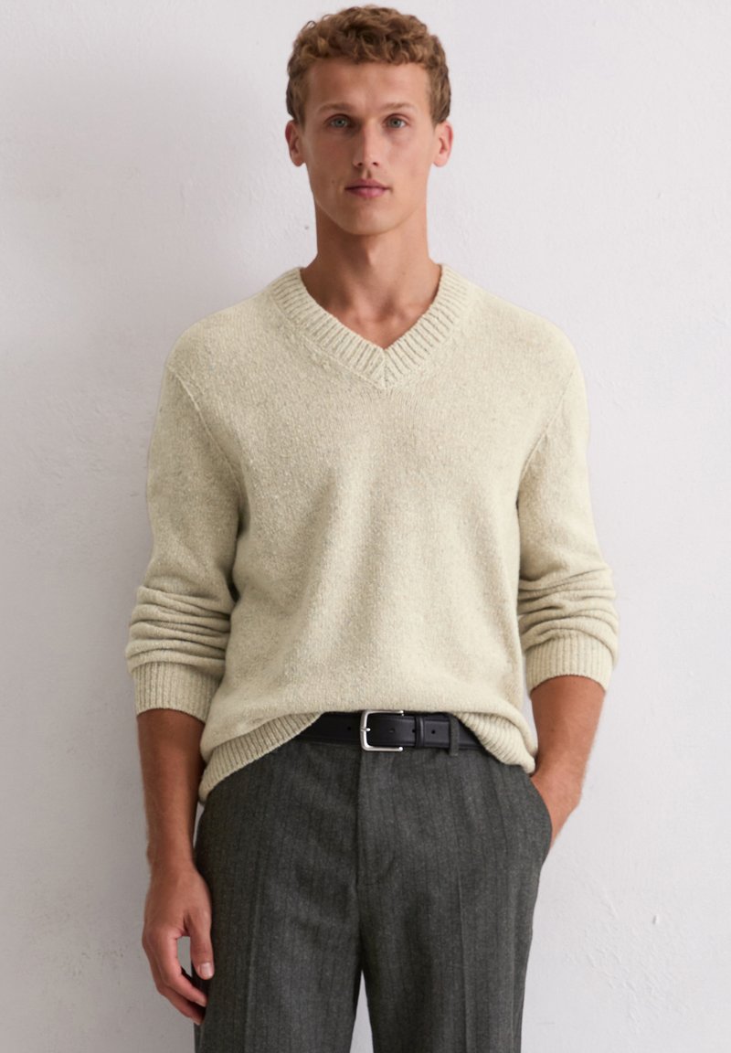 Cremfarbener V-Ausschnitt-Pullover mit strukturiertem Strick, gerippten Bündchen, kombiniert mit grauen Nadelstreifenhosen und einem schwarzen Gürtel. Das Model steht vor einem neutralen Hintergrund.