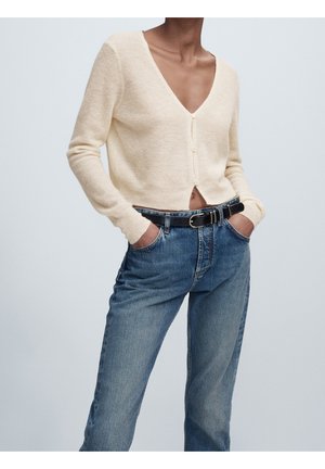 Persona che indossa un cardigan crema con scollo a v e bottoni, infilato leggermente in un paio di jeans a vita alta blu con cintura nera, con le mani nelle tasche.