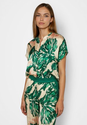 Young woman wearing matching beige outfit with large green and pink floral print, standing against white background.