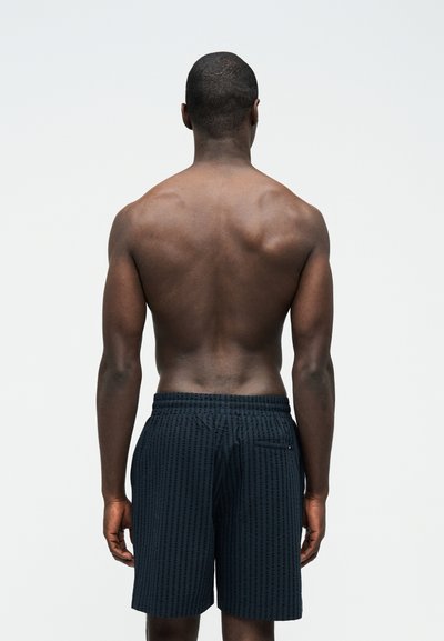 Hombre de piel oscura y cabello corto visto desde atrás, vistiendo pantalones cortos negros con textura sobre un fondo blanco liso.