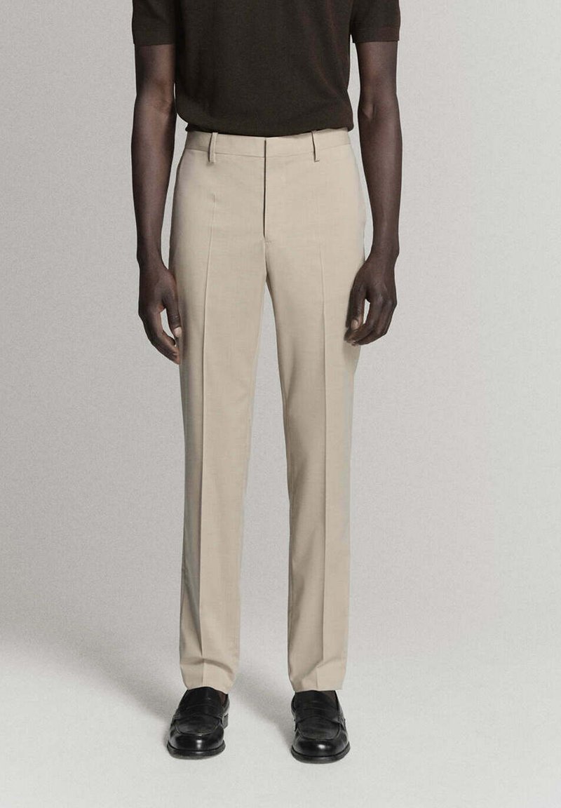 Homme portant un pantalon beige sur mesure, des chaussures noires et un haut à manches courtes foncé, debout devant un fond clair uni.