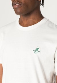 Hvid bomuld t-shirt med rund hals. Har en grøn broderet hajfinne og bølgede linjer på venstre bryst. Glat tekstur.
