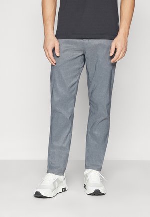 Mann trägt graue Hose, weiße Sneakers und ein schwarzes Shirt und steht vor einem schlichten hellen Hintergrund.