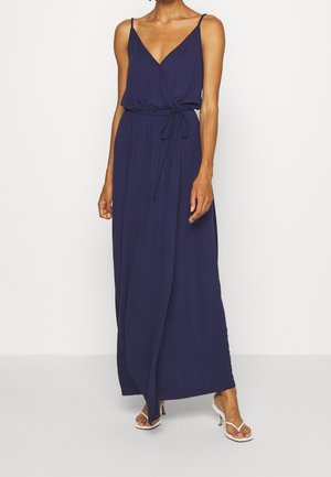 Vrouw draagt een mouwloze marineblauwe maxi-jurk met een V-hals en een taillekoord, gecombineerd met witte sandalen met hak, staand tegen een effen achtergrond.