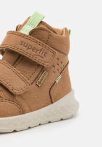 Chaussure en cuir marron avec une finition texturée, bande Velcro, onglet d'accent vert et semelle blanche rembourrée. Présente un marquage Gore-Tex.