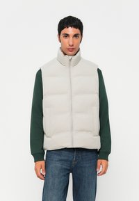Lys grå polstret vest med høj krave, frontlynlås og sidelommer, båret over en mørkegrøn langærmet skjorte og blå jeans.