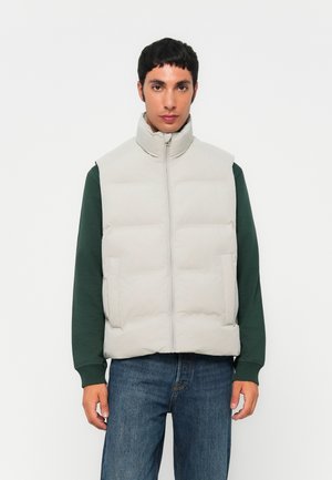 Lichtgrijze gewatteerde vest met hoge kraag, voorrits en zijzakken, gedragen over een donkergroene lange mouwen shirt en blauwe jeans.