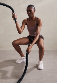 Atletische vrouw in een lichtpaarse tanktop en gestreepte shorts, die een dikke zwarte battle rope hanteert in een squatpositie. Lichte sneakers.