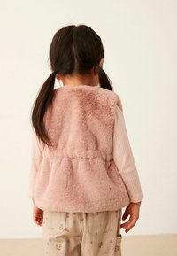 Gilet en fausse fourrure rose pâle, avec une texture douce et une taille cintrée. La tenue est complétée par un pantalon beige à motifs.