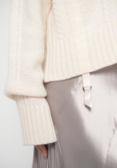 Krémový pletený svetr s žebrovanými manžetami a texturou copánkového pletení, doplněný elegantní bledou saténovou sukní s detailem saténové stuhy.