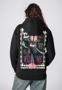 Zwarte hoodie met een kleurrijke circusachtige graphic op de rug, met een personage in een hoge hoed, teksten en geometrische patronen.