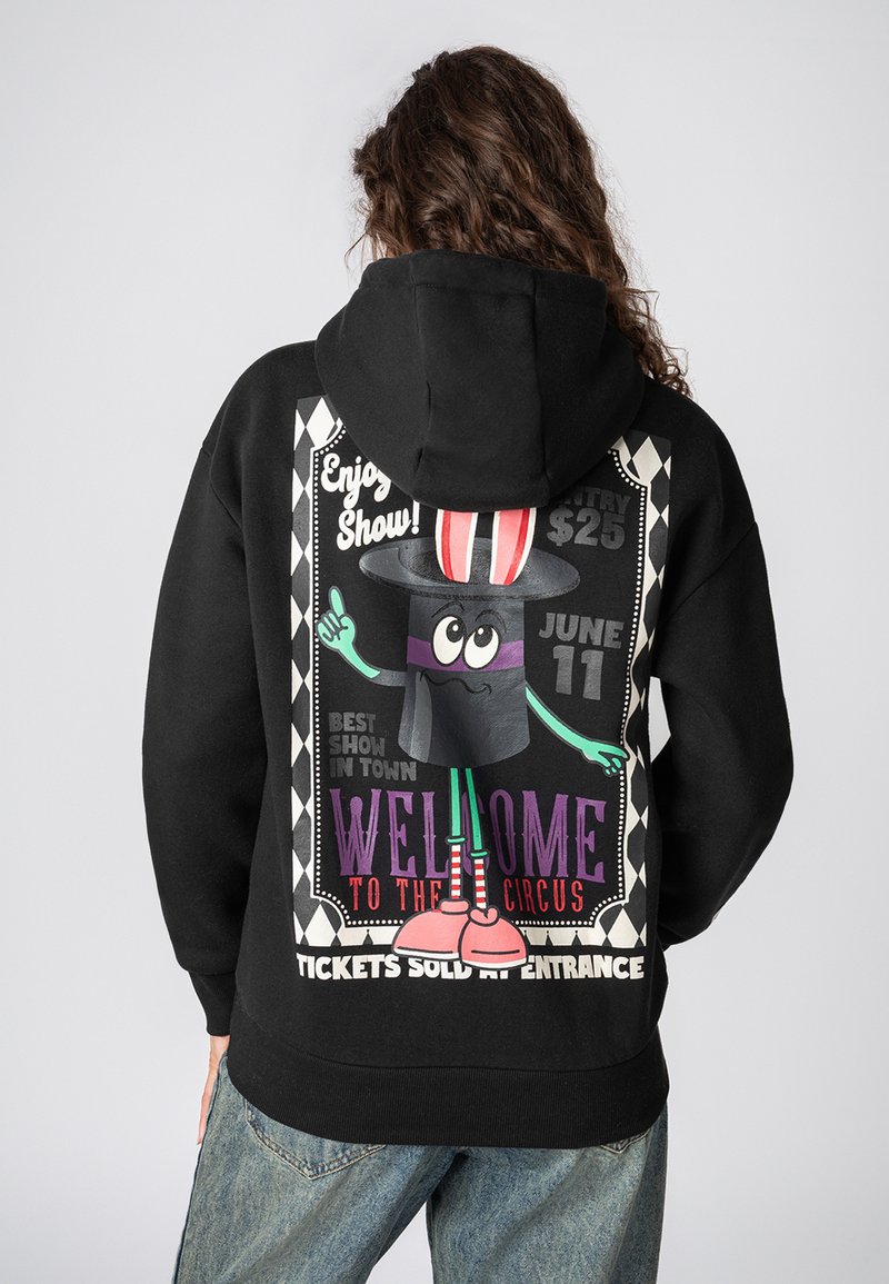 Zwarte hoodie met een kleurrijke circusachtige graphic op de rug, met een personage in een hoge hoed, teksten en geometrische patronen.