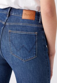 Jeans de mezclilla azul con un diseño ajustado, que presentan un parche de cuero marrón con el logo y un patrón de costura distintivo en el bolsillo trasero.