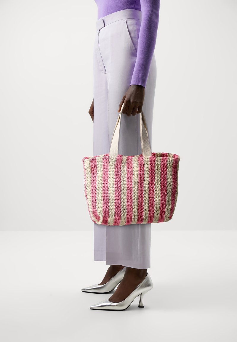 Personne portant un pantalon et un haut lavande tenant un sac cabas tissé rayé rose et beige, portant des chaussures à talons hauts argentées brillantes.