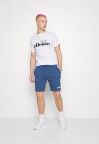 Biely tričko s čiernym logom "ellesse", v kombinácii s Modrými šortkami s šnúrkou a malým bielym logom na leme. Biely tenisky.