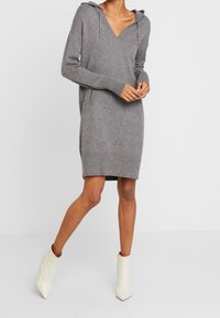 Grijze gebreide hoodie jurk met een V-hals, lange mouwen en zijzakken, gecombineerd met witte enkellaarsjes. Zachte textuur en een ontspannen pasvorm.