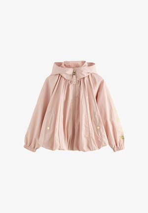 Junges Mädchen mit langen blonden Haaren trägt eine hellrosa Kapuzenjacke, schwarze Hose und weiße Turnschuhe, steht mit einer Hand in der Tasche.