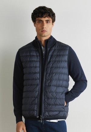 Uomo con capelli scuri e ricci che indossa un gilet trapuntato blu navy sopra una maglia a maniche lunghe scura, in piedi contro un muro chiaro e semplice.