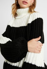 Gruby sweter z golfem w czarno-białe paski, z fakturą rib i oversized rękawami, zapewniającymi dodatkowe ciepło.